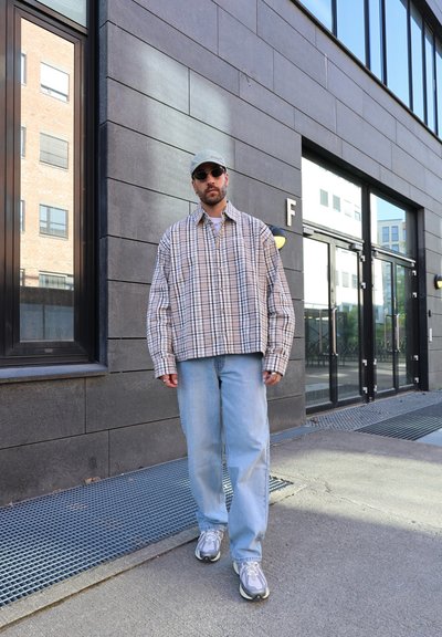 Camisa de franela beige de gran tamaño y a cuadros, jeans de mezclilla azul claro, zapatillas grises con detalles en blanco y una gorra clara. Gafas de sol oscuras.