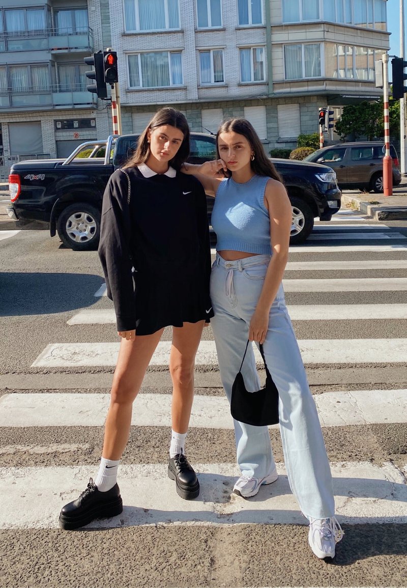 To personer poserer på et fodgængerfelt. Den ene har en sort oversized sweatshirt og nederdel på, mens den anden har en lys blå crop top med høj talje jeans.