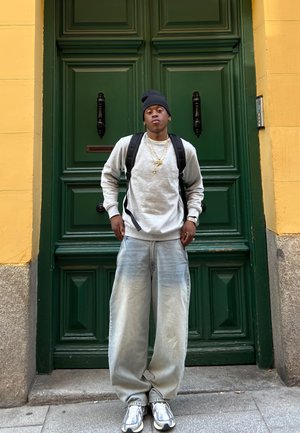 Sudadera gris, jeans baggy azul claro y zapatillas plateadas. Mochila negra y gorro de lana. De pie frente a una puerta verde oscura.