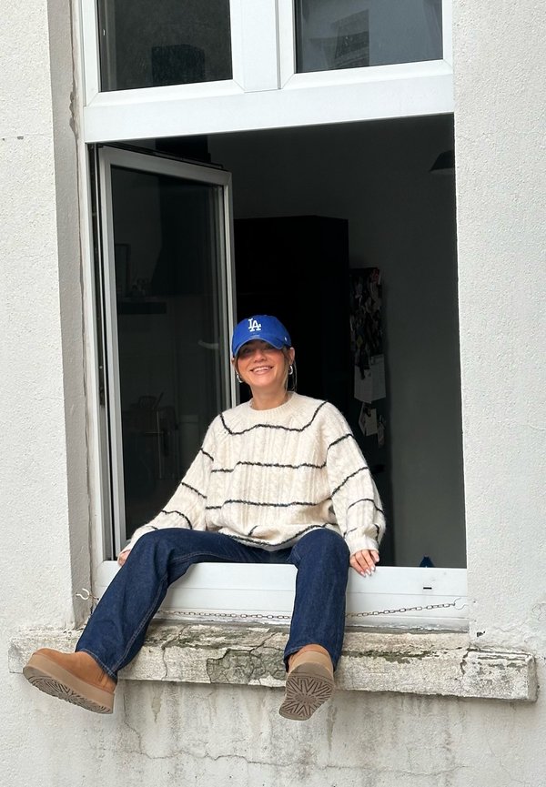 Valkoinen neulottu villapaita, jossa on mustia raitoja, sininen baseball-hattu, farkut ja vaaleat saappaat, istuu ikkunalaudalla.