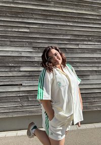 Balta Adidas drēbe ar zaļām svītrām, kas ietver īsām piedurknēm t-kreklu un šortus. Iespējas ietver priekšējo kabatu un logo akcentus. Koksnes fons.