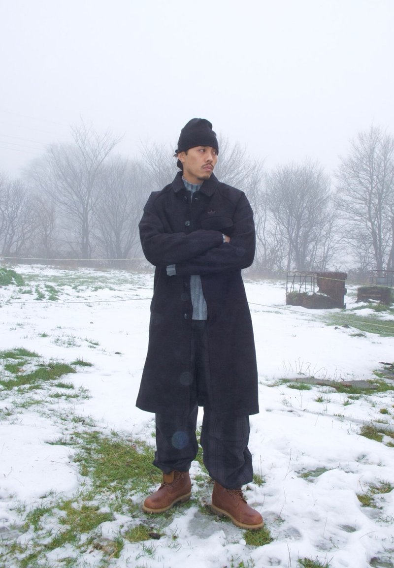 Homme portant un manteau noir, un bonnet et des bottes brunes, debout les bras croisés sur une herbe enneigée avec des arbres nus dans un paysage hivernal brumeux.
