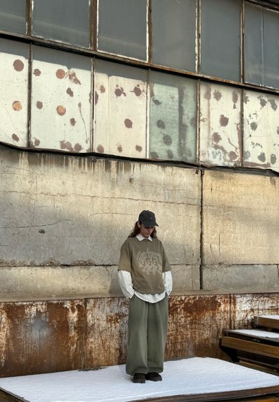 Camisa verde oliva de gran tamaño con diseño gráfico sobre una camisa blanca a rayas, combinada con pantalones verdes holgados y zapatos oscuros frente a un fondo industrial.