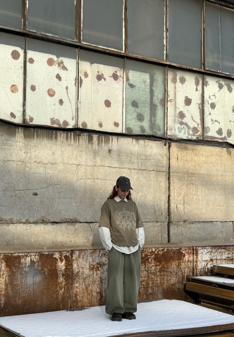 Chemise oversize vert olive avec motif graphique portée sur une chemise blanche à rayures, associée à un pantalon large vert et des chaussures foncées, sur un fond industriel.