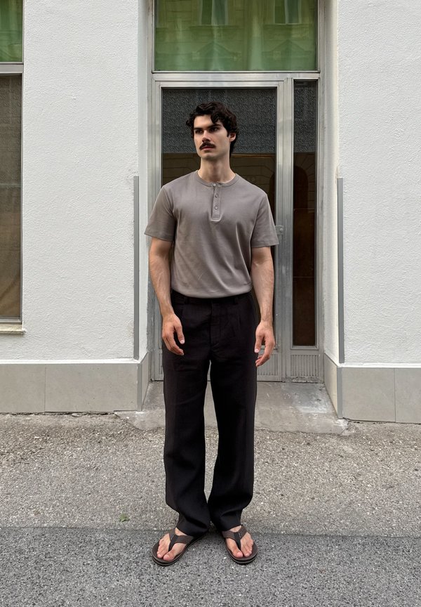 Mand med mørkt hår og overskæg iført en grå henley t-shirt med korte ærmer, sorte bukser og brune sandaler, står udenfor en hvid bygningsdør.