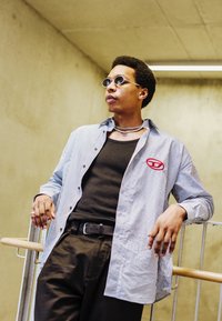 Camicia a righe blu e bianca, indossata sopra una canotta nera. Gli accessori includono occhiali da sole e collane d'argento sovrapposte.