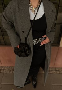 Šedý vlněný kabát, priliehavé čierne šaty, opasok so striebornou kovovou prackou s kruhovými detailmi, čierna kožená kabelka, dlhé nechty, čierne špicaté čižmy.