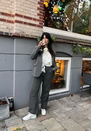 Traje gris con una chaqueta ajustada y pantalones de pierna ancha, combinado con una blusa blanca con volantes. Sosteniendo una copa de vino, de pie sobre un pavimento de piedra.