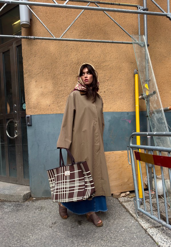 Mujer con abrigo beige y pañuelo floral en la cabeza lleva una gran bolsa a cuadros marrón y blanca, de pie en una calle cerca de andamios y un edificio.