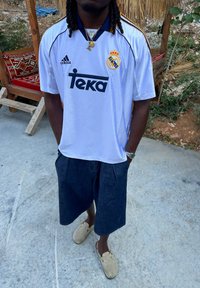 Balta futbolo marškiniai su juodais ir auksiniais akcentais, mėlynos džinsinės šortai ir kreminiai mediniai šlepetės. Papuošalai apima grandines ir pakabuką.