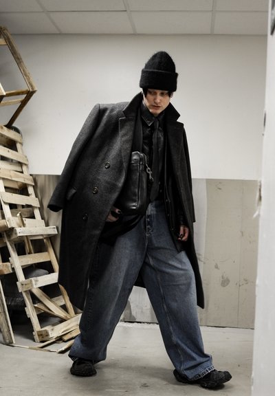 Abrigo gris de lana oversize con botones cruzados, bolso negro de cuero, camiseta negra y vaqueros azules holgados. Gorro oscuro y zapatos gruesos.