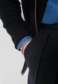 Personne portant un pantalon et une veste noirs avec un poignet de chemise bleu visible, main dans la poche du pantalon, et un bracelet chaîne argenté au poignet.