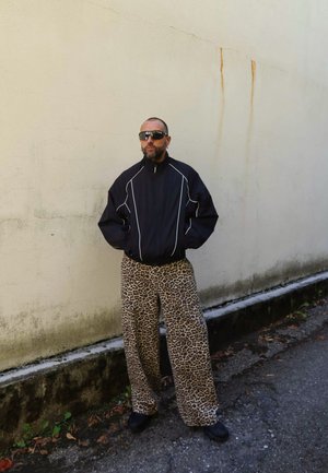 Hombre con barba y cabeza rapada, vestido con chaqueta negra, gafas de sol y pantalones de leopardo, de pie contra una pared manchada beige al aire libre.