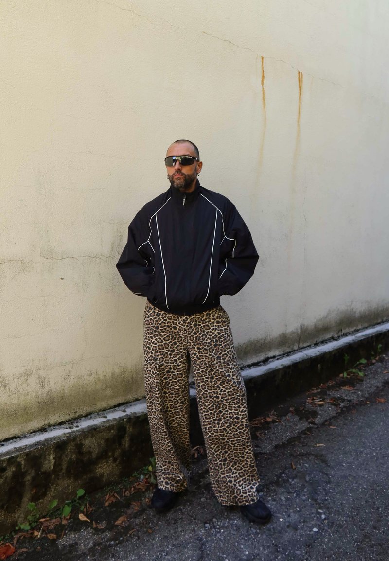 Uomo con barba e testa rasata che indossa una giacca nera, occhiali da sole e pantaloni a stampa leopardata, in piedi contro un muro beige macchiato all'aperto.