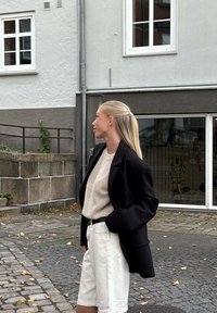 Czarna, oversized marynarka, beżowy sweter dzianinowy i białe szorty. Gładkie tekstury, luźny krój, zaprezentowane na tle brukowanej ulicy.