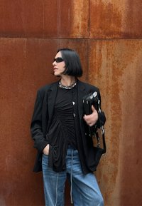 Blazer preto sobre uma blusa preta franzida, jeans azul de corte largo, colar de corrente prateada e óculos de sol escuros, com um fundo de cor ferrugem.