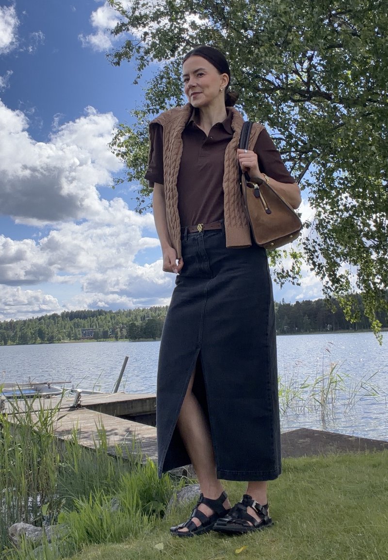 Femme debout près d’un lac, portant une chemise marron, un gilet tricoté, une longue jupe sombre fendue devant, des sandales noires, et tenant un sac à main marron.