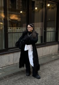 Sort faux fur-frakke med en struktureret krave, lagdelt over en sort top og hvid taljevikle. Sorte bukser og chunky fodtøj fuldender outfittet.