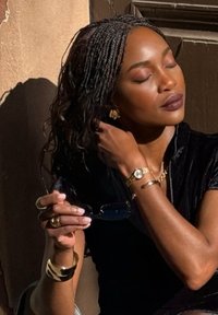 Accesoriile includ bijuterii din aur: o brățară mare, un ceas cu cadran rotund și cercei cu tortiță. Silueta poartă un top negru și ține ochelari de soare.