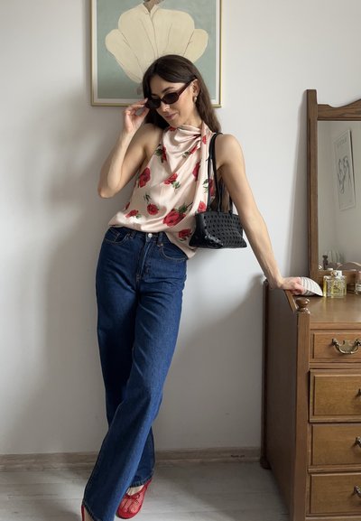 Top sin mangas de flores en crema con rosas rojas, combinado con jeans de tiro alto azul y zapatos rojos. Sosteniendo un pequeño bolso negro y gafas de sol.