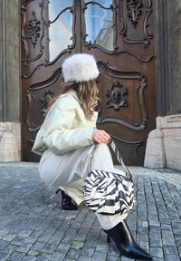 Inimene helerohelises riietuses kummardub, hoides musta-valgete triipudega kotti, taustal kaunis puidust uks. Silma paistavad mustad kontsaga saapad.