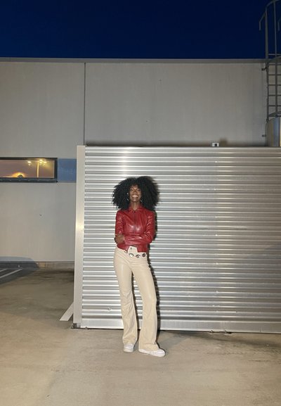 Chaqueta de cuero roja, pantalones acampanados beiges, zapatillas blancas. De pie contra una pared metálica corrugada, ambiente exterior al anochecer.