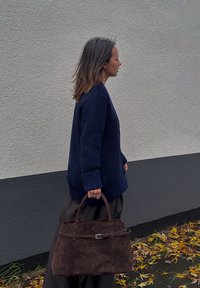 Bolsa de camurça castanha com uma forma estruturada, alças superiores e um detalhe de alça plana. Usada por uma pessoa que veste um suéter canelado azul-marinho e uma saia escura.