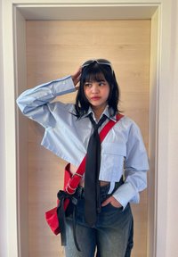 Camicia azzurra a maniche corte con bottoni e cravatta nera, abbinata a pantaloni di jeans scuri. Una borsa a spalla rossa con dettaglio a fiocco completa l'outfit.