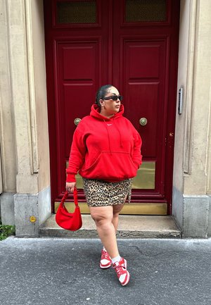 Sudadera roja con bolsillo delantero, falda mini de estampado de leopardo, bolso rojo y zapatillas rojas con acentos blancos. De pie frente a puertas rojas.