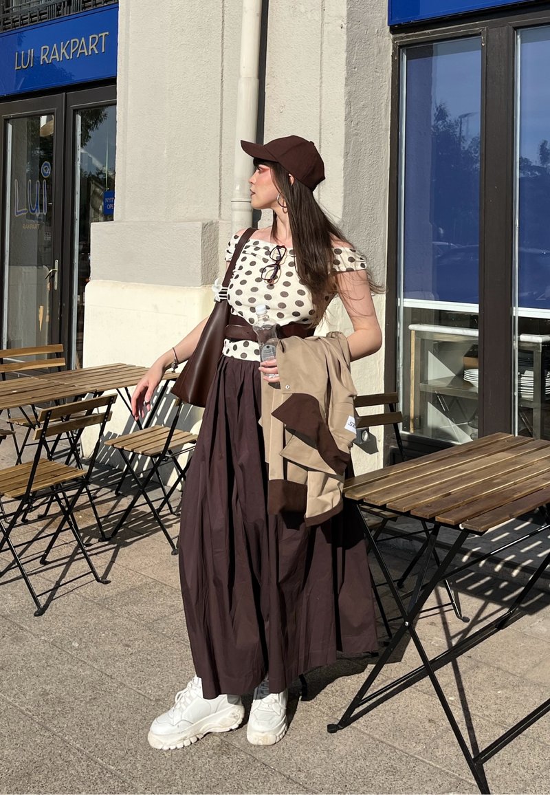 Femme portant une casquette marron, un haut à pois et une longue jupe foncée se tient devant un café, tenant une bouteille d’eau et une veste beige à côté de tables et de chaises en bois.