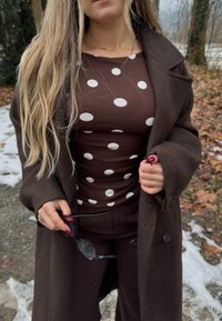 Femme portant un haut marron à pois et un long manteau sombre, tenant des lunettes de soleil noires en plein air, avec de la neige et des feuilles mortes à proximité.