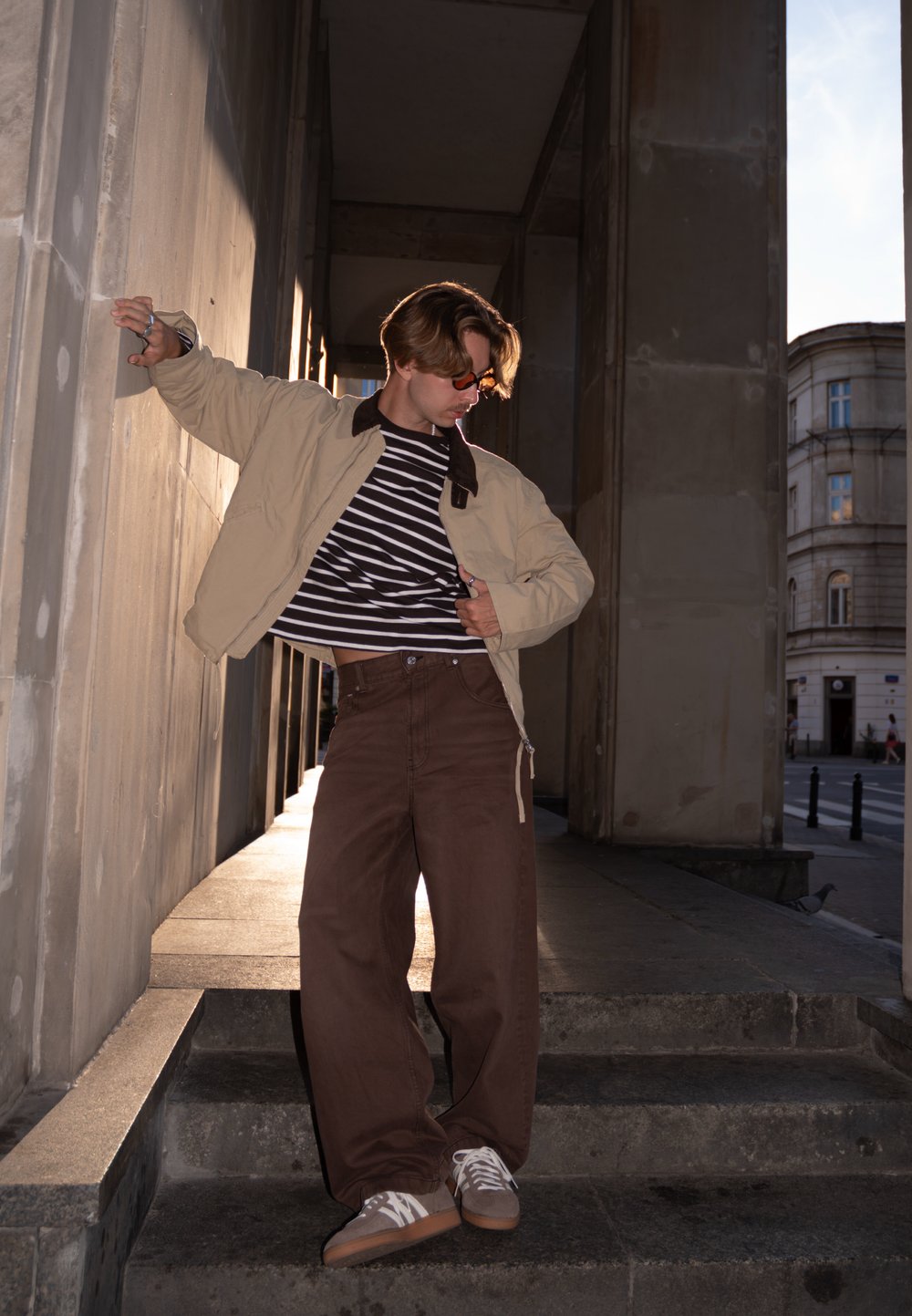 Bēša jaka ar brūnu apkakli, melni balta svītraina krekls, brūnas plašas bikses un pelēki sporta apavi uz akmens kāpnēm.