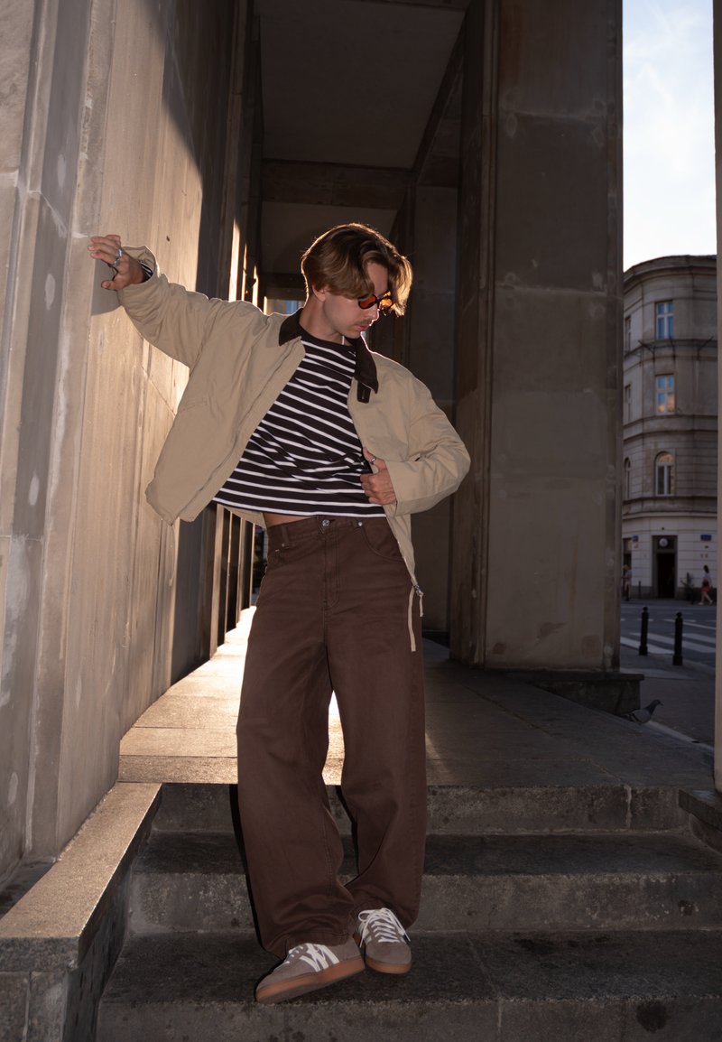 Bēša jaka ar brūnu apkakli, melni balta svītraina krekls, brūnas plašas bikses un pelēki sporta apavi uz akmens kāpnēm.