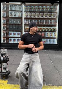 Fekete testhezálló póló, laza szürke farmernadr�ág fakó mosással, ezüst ékszerek és fülhallgató. A háttérben különféle magazinok láthatók egy kijelzőn.