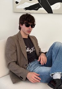 Blazer a motivo houndstooth marrone sopra una t-shirt grafica nera, abbinato a jeans strappati azzurri chiari e scarpe nere, seduto su un divano chiaro.