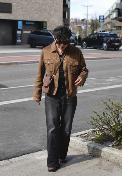 Persona con chaqueta marrón, pantalones negros y gafas de sol caminando por una acera urbana cerca de coches aparcados y un pequeño arbusto.
