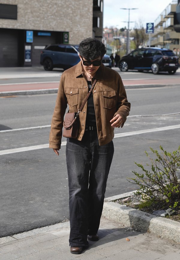 Person iført brun jakke, sorte bukser og solbriller går på en urbane fortov nær parkerede biler og en lille busk.