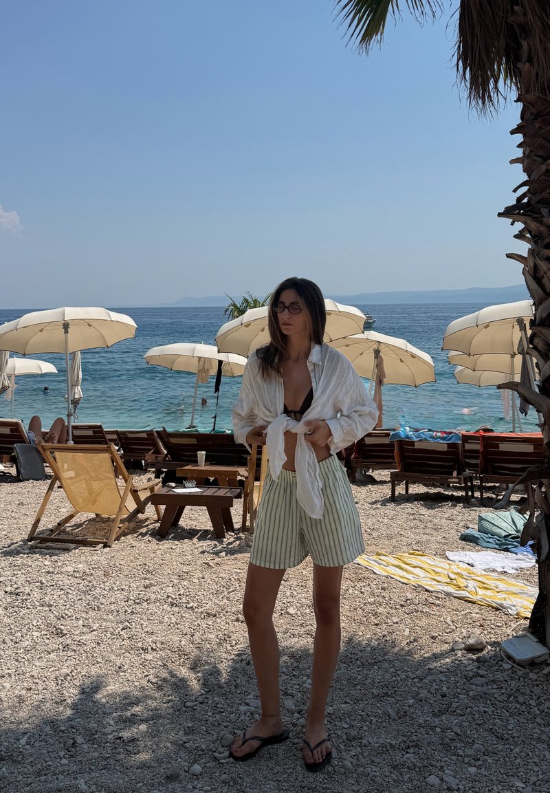 Femme en lunettes de soleil et short rayé se tient sur une plage de galets près de parasols et de chaises longues au bord de la mer sous un ciel clair.