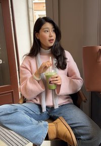 Mujer joven con suéter rosa y jeans sentada en el interior, sosteniendo una bebida fría verde con una pajita, mirando hacia un lado, con botas color bronce y una bolsa cerca.