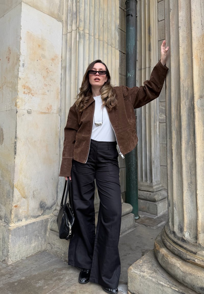 Femme portant une veste marron, une chemise blanche, un pantalon large noir et des lunettes de soleil, debout près de colonnes en pierre, tenant un sac à main noir.