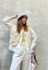Beige knitted vest with a patterned design over a light green top, teamed with white trousers adorned with floral embroidery and a pastel bucket hat.