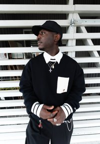 Homme portant une casquette noire, un pull noir avec col et poche blancs, des colliers en couches, des bagues et une ceinture chaîne, debout devant un mur blanc à lattes.