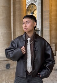 Fiatal személy túlságosan nagy fekete bőrdzsekit, barna inget, világos mintás nyakkendőt és ezüst láncnyakláncot viselve, kőoszlopok mellett áll.