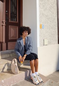 Blazer cinza sobre uma blusa preta, saia preta e ténis bege com detalhes coloridos. Segurando uma bebida, sentado em degraus de pedra.