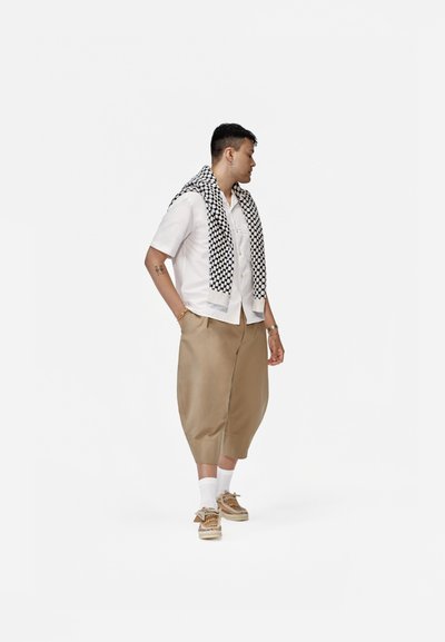Camisa blanca de manga corta, pantalones cortos beige holgados, bufanda negra y blanca con estampado, y zapatos estampados, con calcetines blancos.