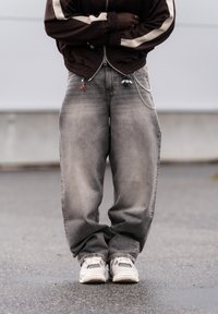 Personne portant un jean gris délavé surdimensionné avec des chaînes et des breloques, des baskets blanches, et une veste marron avec des bandes blanches, debout à l'extérieur.