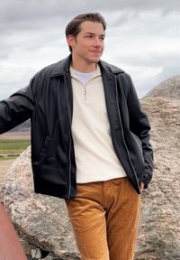 Svart jacka med dragkedja framtill, med en slät yta, över en ljus beige zip-längd tröja, matchad med bruna kordflöjlsbyxor. Klippig utomhusmiljö.