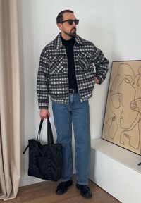 Svart-hvitt rutete jakke med to frontlommer, svart turtleneck, blå denimjeans, svart handlenett og mørke solbriller.
