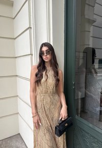 Mujer con vestido sin mangas con patrón dorado y gafas de sol oscuras está contra una pared blanca sosteniendo un bolso negro cerca de una puerta de cristal.