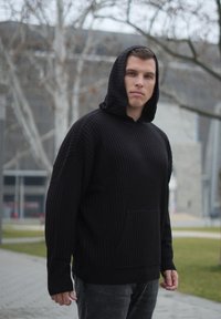 Sweat à capuche noir côtelé avec une poche avant, des manches longues et une capuche avec cordon de serrage. Porté avec un jean foncé dans un cadre extérieur.